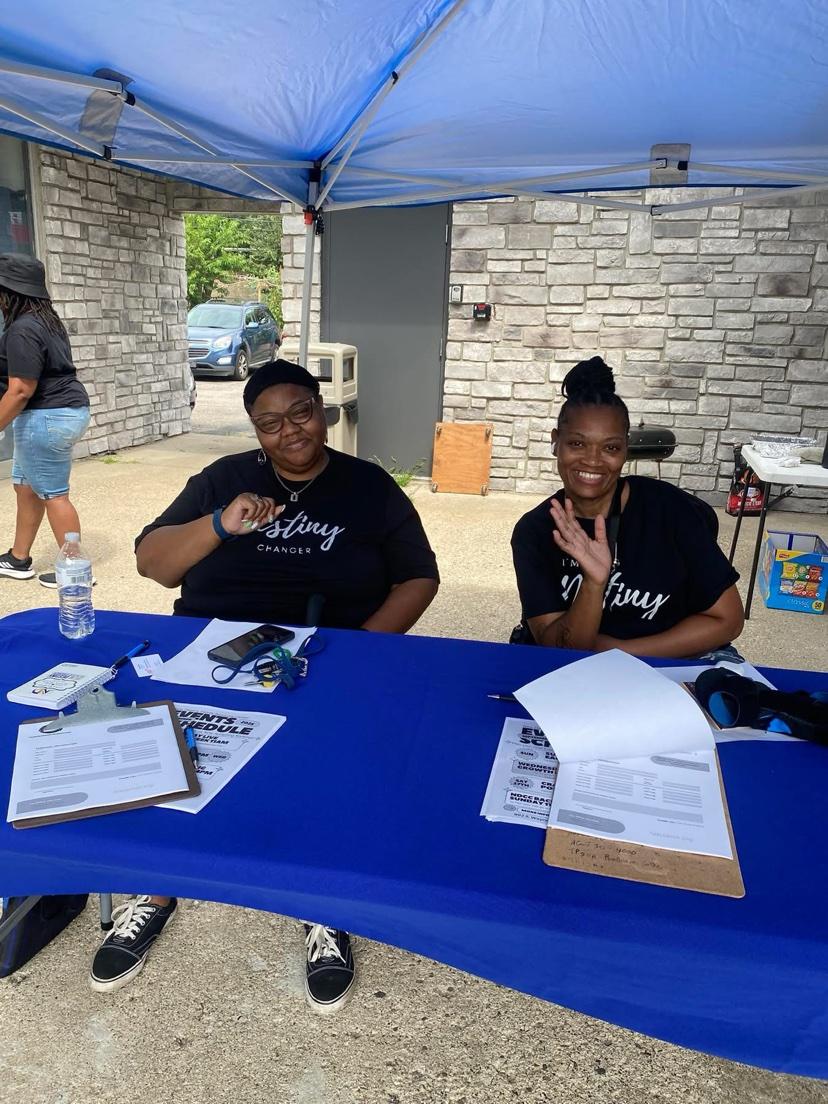 Two volunteers wearing “I'm a Destiny Changer” shirts at a welcome table under a tent